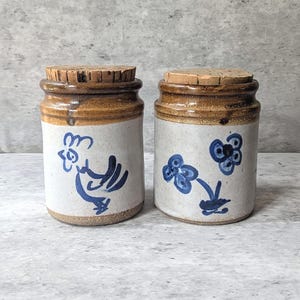 May include: Two cylindrical ceramic jars with cork lids. The jars have a light grey base with a brown glazed rim. One jar features a blue hand-painted rooster, while the other jar displays a blue hand-painted floral design on the front surface.
