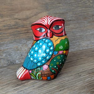 May include: A hand-painted wooden owl figurine. The owl has a red head with white stripes, black eyes, and a blue wing with white polka dots. The body is decorated with trees, flowers, and other colourful details. The owl is on a wooden surface.