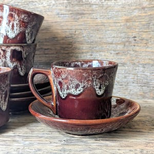 May include: A set of vintage ceramic coffee cups and saucers. The cups and saucers are a deep burgundy colour with a white, textured glaze around the rim. The cups have a handle and sit on a matching saucer. A stack of bowls is visible in the background.