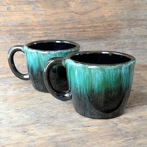 May include: Two ceramic mugs with a teal and black glaze. The mugs have a rounded shape with a black handle and interior. The glaze creates a dripping effect, with the teal colour fading into black at the base. The mugs are on a wooden surface.