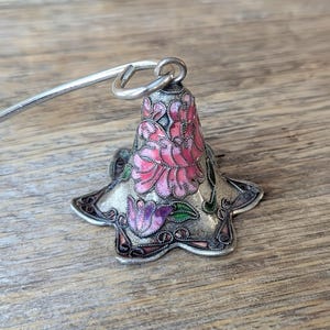 May include: A silver-coloured bell-shaped ornament with a floral design. The bell features pink and purple flowers with green leaves, set against a silver background. A silver handle is attached to the top. The ornament is likely for decorative purposes.