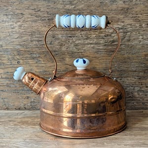 May include: A copper kettle with a white and blue ceramic handle and spout. The kettle has a round body and a lid with a matching ceramic knob. The copper has a warm, reflective finish. The handle is curved.