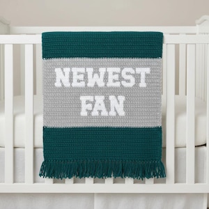May include: A teal, grey, and white crochet baby blanket with the words "NEWEST FAN" in white. The blanket is draped over a cot. The bottom edge has a fringe. The blanket is handmade.