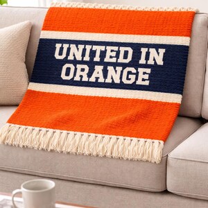 May include: Orange and navy blue throw blanket with the text "UNITED IN ORANGE" in white block letters. The blanket has a cream-colored fringe along the bottom edge and is draped over a gray sofa.
