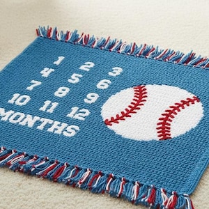 May include: A blue crocheted baby blanket with a baseball theme. The blanket features a white baseball with red stitching and the numbers 1-12, with the word "MONTHS". The blanket has a fringe border with red, white, and blue accents.