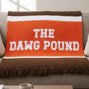 May include: A handmade crochet blanket in orange, white, and brown, with the text "THE DAWG POUND" in white block letters. The blanket has a fringed edge and is draped over a beige sofa. A coffee table with a mug and books is in the foreground.