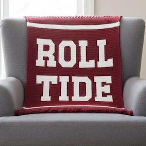 Pode incluir: Uma manta de crochê bordô e branca drapeada sobre uma poltrona cinza. A manta apresenta as palavras "ROLL TIDE" em letras brancas grandes. A manta tem uma borda branca e franjas.