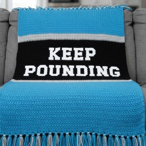 May include: A blue, black, and gray crocheted throw blanket with the words "KEEP POUNDING" in white block letters. The blanket has a fringe along the bottom edge and is draped over a gray couch.