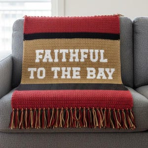 May include: A crocheted throw blanket in red, black, and tan, draped over a gray sofa. The blanket features the text "FAITHFUL TO THE BAY" in white block letters and has a fringe along the bottom edge.