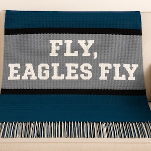 May include: A knitted blanket in shades of blue, gray, and black, draped over a beige sofa. The blanket features the text "FLY, EAGLES FLY" in white block letters. The bottom edge has a fringe.