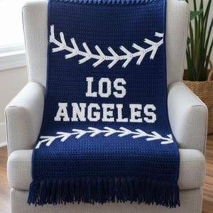 May include: A navy blue crocheted blanket draped over a gray armchair. The blanket features white baseball stitching designs and the words "LOS ANGELES" in bold white letters. The bottom edge has a fringe.
