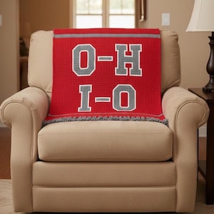 May include: A red throw blanket with the letters "O-H I-O" in gray and white, draped over a beige armchair. The blanket has a gray border and fringe, and the letters are block-style.