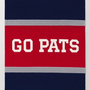 May include: A rectangular, knitted blanket in navy blue, red, and gray, with a fringe along the bottom edge. The words "GO PATS" are displayed in large, white, block letters across the center red stripe.