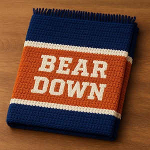以下が含まれることがあります： 大きな白い文字で「BEAR DOWN」と書かれた、ネイビー、オレンジ、白の編み物ブランケット。 ブランケットにはフリンジの縁があり、きれいに折りたたまれています。