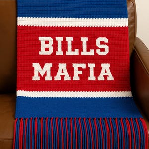 Op de afbeelding: Een gebreide plaid in blauw, rood en wit met de tekst "BILLS MAFIA" in witte letters. De deken heeft een franje van blauw en rood garen en is gedrapeerd over een bruine leren bank.