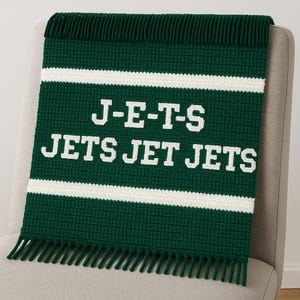 Könnte beinhalten: Eine grün-weiße Häkeldecke mit dem Text "J-E-T-S JETS JET JETS" in Weiß. Die Decke hat weiße Streifen und einen Fransenbesatz am unteren Rand. Die Decke ist über einen grauen Stuhl drapiert.