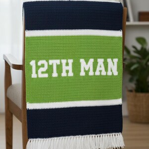 May include: A navy blue, green, and white crocheted blanket draped over a wooden chair. The blanket features the text "12TH MAN" in white block letters on a green stripe. White fringe trims the bottom.