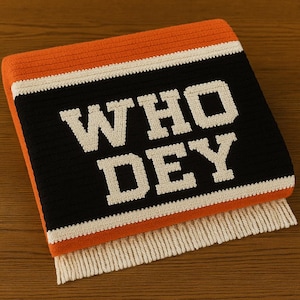 Könnte beinhalten: Eine orange, schwarz und weiß gestrickte Decke mit dem Text "WHO DEY" in fetten, weißen Buchstaben. Die Decke hat einen Fransenrand und ist gefaltet. Das Farbschema ist orange, schwarz und weiß.