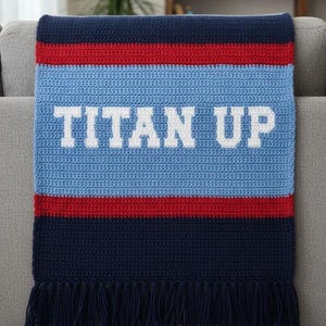 May include: A crocheted blanket in navy blue, red, and light blue, with the words "TITAN UP" in white block letters. The blanket has a fringed edge and is draped over a gray sofa.