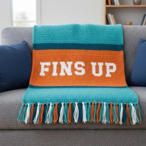 May include: A turquoise, orange, and teal crocheted blanket with the words "FINS UP" in white. The blanket has a fringe of teal, orange, and white tassels. The blanket is draped over a gray couch with blue pillows.
