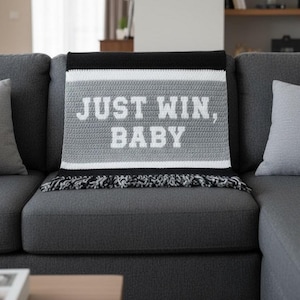 May include: A crocheted throw blanket in shades of gray, black, and white, draped over a gray sofa. The blanket features the text "JUST WIN, BABY" in white block letters. The bottom edge has a fringe.