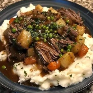 Puede incluir: Un plato de estofado con puré de patatas. El plato incluye carne de res tierna y desmenuzada, zanahorias, patatas y guisantes en una salsa rica, adornado con hierbas frescas. Servido en un plato azul oscuro, esta comida es un plato reconfortante clásico.