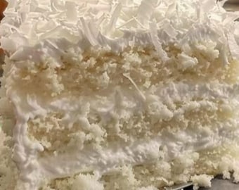 May include: A slice of coconut cake with multiple layers of white frosting and shredded coconut topping. The cake has a moist appearance, with visible layers of cake and frosting. The dessert is presented on a silver surface.