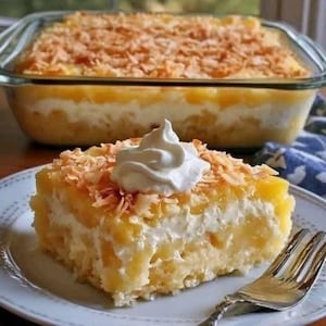 May include: A slice of layered dessert on a white plate, topped with whipped cream and toasted coconut flakes. The dessert has a yellow and white filling. A glass baking dish with the same dessert is in the background.