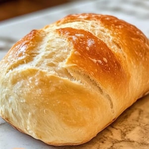 Puede incluir: Un pan recién horneado con una corteza dorada y un interior ligero y aireado. El pan tiene forma rectangular con la parte superior ligeramente redondeada y cortes visibles. La corteza es de un color cálido y rico, y el pan parece suave.