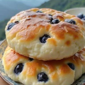 Può includere: Una pila di due focaccine ai mirtilli dorate. I prodotti da forno hanno un interno morbido e cremoso e sono cosparsi di mirtilli freschi, blu scuro. Le focaccine sono su un piatto decorativo, con uno sfondo montuoso sfocato.