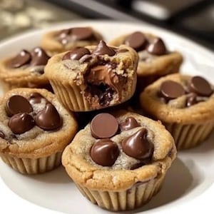 May include: A plate of mini chocolate chip cookie cups. The golden-brown baked cups are filled with chocolate and topped with chocolate chips. One cookie cup has a bite taken out of it, revealing the chocolate filling.