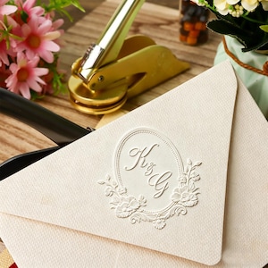 Op de afbeelding: Ivoorkleurige envelop met een reliëf bloemmotief en de initialen "K & G". Een gouden embossing tool is zichtbaar, wat duidt op trouwbriefpapier. Roze bloemen en een houten oppervlak completeren de scène.