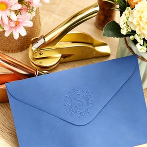 Puede incluir: Un sobre azul que presenta un escudo en relieve en el centro con la letra S. Detrás del sobre se encuentra una herramienta de estampado de metal dorado con un mango curvo y una base circular, utilizada para crear impresiones en papel.
