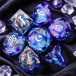 May include: A set of seven polyhedral dice featuring a deep blue and purple galaxy-inspired resin base with embedded gold leaf flakes. Each die has sharp edges and is engraved with silver-colored numbers in a gothic font, with a dragon icon.