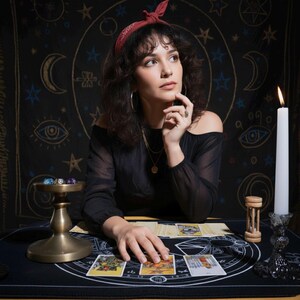 Puede incluir: Una mujer con una diadema roja y un top negro con hombros descubiertos est&aacute; sentada en una mesa con cartas del tarot. Una vela encendida, un reloj de arena y un cuenco de lat&oacute;n con dados tambi&eacute;n est&aacute;n sobre la mesa. El fondo es un tapiz negro con s&iacute;mbolos celestiales.