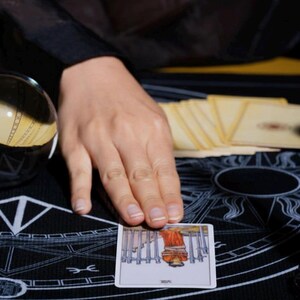 Puede incluir: Una carta de tarot con una figura con t&uacute;nica roja y espadas plateadas se muestra sobre una estera negra con s&iacute;mbolos blancos. Tambi&eacute;n se ven una bola de cristal y una baraja de cartas. Una mano toca la carta.