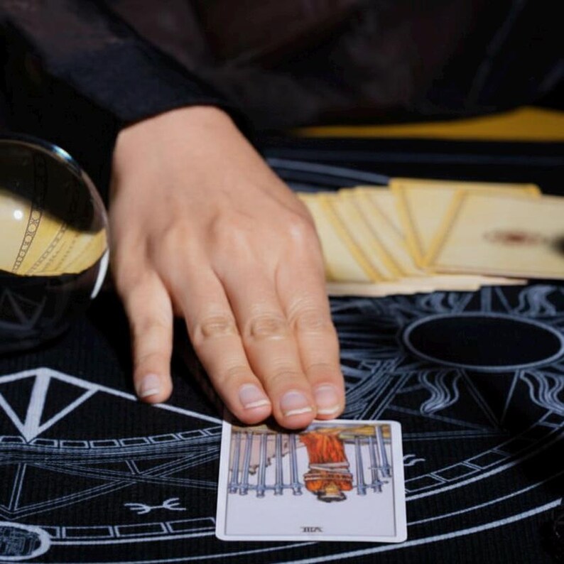 Puede incluir: Una carta de tarot con una figura con t&uacute;nica roja y espadas plateadas se muestra sobre una estera negra con s&iacute;mbolos blancos. Tambi&eacute;n se ven una bola de cristal y una baraja de cartas. Una mano toca la carta.