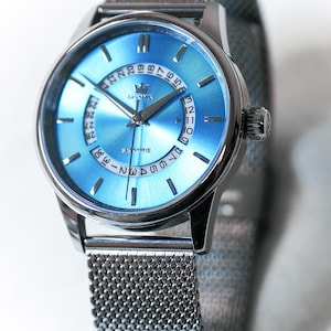 May include: A silver wristwatch with a light blue face and a mesh strap. The watch face displays the date and time with silver hands and markers. The word "SAPPHIRE" is printed on the watch face.