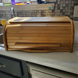 May include: A bamboo bread box with a roll-top lid. The box is made of light brown bamboo slats and has a curved design. The box is sitting on a white countertop in a kitchen.