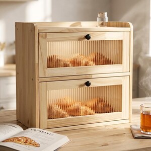 May include: A light-colored wooden bread box with two pull-out drawers, each with a clear, ribbed glass panel. The drawers are filled with croissants. A glass of tea and a cookbook are on the wooden table.