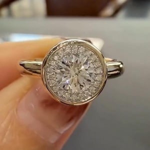 May include: A gold ring with a large, round, clear gemstone at the center, surrounded by a halo of smaller, clear stones. The ring's band is a simple, polished gold. The ring is held up in front of a neutral background.