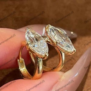 May include: A pair of gold hoop earrings, each featuring a marquise-cut, clear gemstone. The earrings have a polished finish and are set in a gold metal. The image is a close-up, highlighting the sparkle of the gemstones.