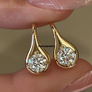 May include: A pair of gold drop earrings, each featuring a single round brilliant-cut clear gemstone set within a smooth, polished teardrop-shaped metal frame. The text DiamoraJewelUS is visible between the two earrings on the gold surface.