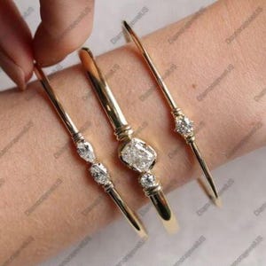 May include: Three gold bangles with diamond accents. The bangles are thin and delicate, with a central diamond setting and smaller diamonds on the other bangles. The diamonds are clear and sparkling, set in a gold band.