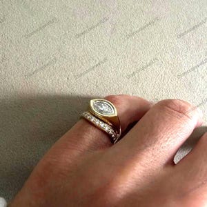 May include: A gold ring with a marquise-cut diamond center stone. The ring is set in a bezel setting and is accompanied by a smaller band with pave-set diamonds. The rings are on a finger.