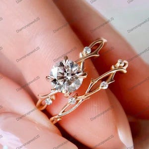 May include: A gold-colored engagement ring set featuring a large, round, clear gemstone in a prong setting. The band has a nature-inspired design with small accent stones. The ring is shown on a person's finger.