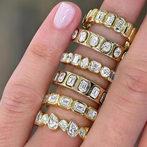 May include: A collection of gold rings with various diamond shapes, including emerald, round, and pear cuts. The rings are displayed on a finger, showcasing their different designs and settings. The rings have a polished, shiny finish.