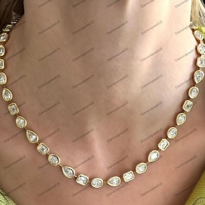 Puede incluir: Un collar de oro con una serie de diamantes engarzados en bisel en varias formas, incluyendo pera, ovalada y rectangular. Los diamantes son claros y brillantes, engarzados en una delicada cadena de oro. El collar se muestra en el cuello de una persona.