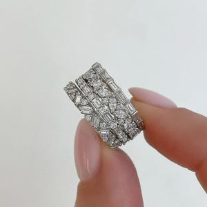 May include: Four silver rings with various diamond cuts, including baguette, marquise, and round stones. The rings are stacked together, showcasing the different designs and sparkle. The jewelry is held between fingers.