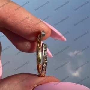 May include: A gold-coloured hoop earring featuring a line of small, rectangular, clear stones. The earring is held between fingers against a light blue backdrop. The earring has a unique, open design.